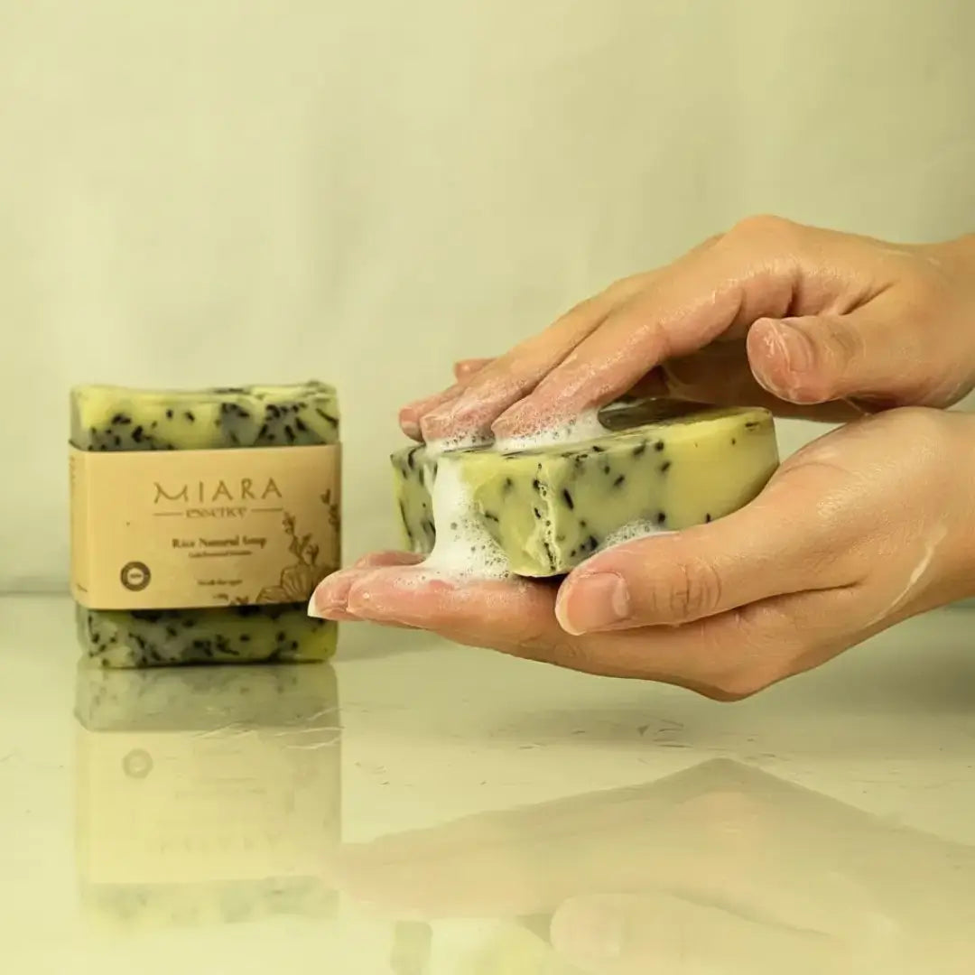 Hand holding a bar of Miara Essence Rice Natural soap with 'MIARA' branding, next to a similar bar on a reflective surface.