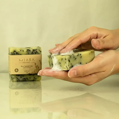 Hand holding a bar of Miara Essence Rice Natural soap with 'MIARA' branding, next to a similar bar on a reflective surface.